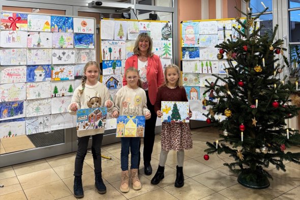 Zu sehen ist Landrätin Anke Beilstein mit den Gewinnerinnen des Malwettbewerbes. Im Hintergrund sind die weiteren eingereichten Bilder abgebildet. Rechts im Bild ist ein Weihnachtsbaum zu sehen. 