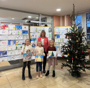 Zu sehen ist Landrätin Anke Beilstein mit den Gewinnerinnen des Malwettbewerbes. Im Hintergrund sind die weiteren eingereichten Bilder abgebildet. Rechts im Bild ist ein Weihnachtsbaum zu sehen. 