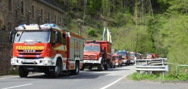 Feuerwehrfahrzeuge fahren eine Straße entlang. Im Hintergrund ist eine Hauswand zu sehen.