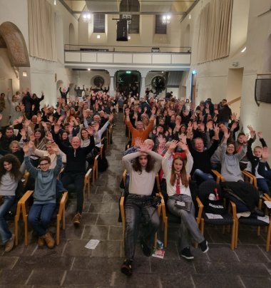 Zu sehen sind viele Kinder und Erwachsene in einem Großen Saal. Sie sitzen reihenweise auf Stühlen.