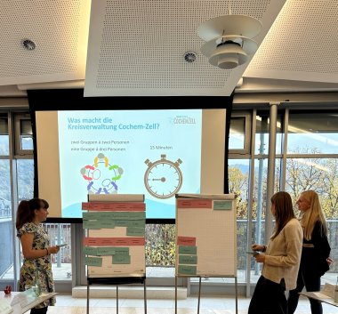 Einführung Zu sehen sind drei Personen die vor zwei Pinnwänden stehen. Im Hintergrund ist eine Leinwand zu sehen, an der eine Präsentation zu sehen ist