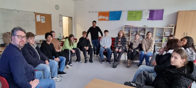 Das Bild zeigt im Stuhlkreis sitzende Schülerinnen und Schüler sowie Lehrkräfte/Dozenten.