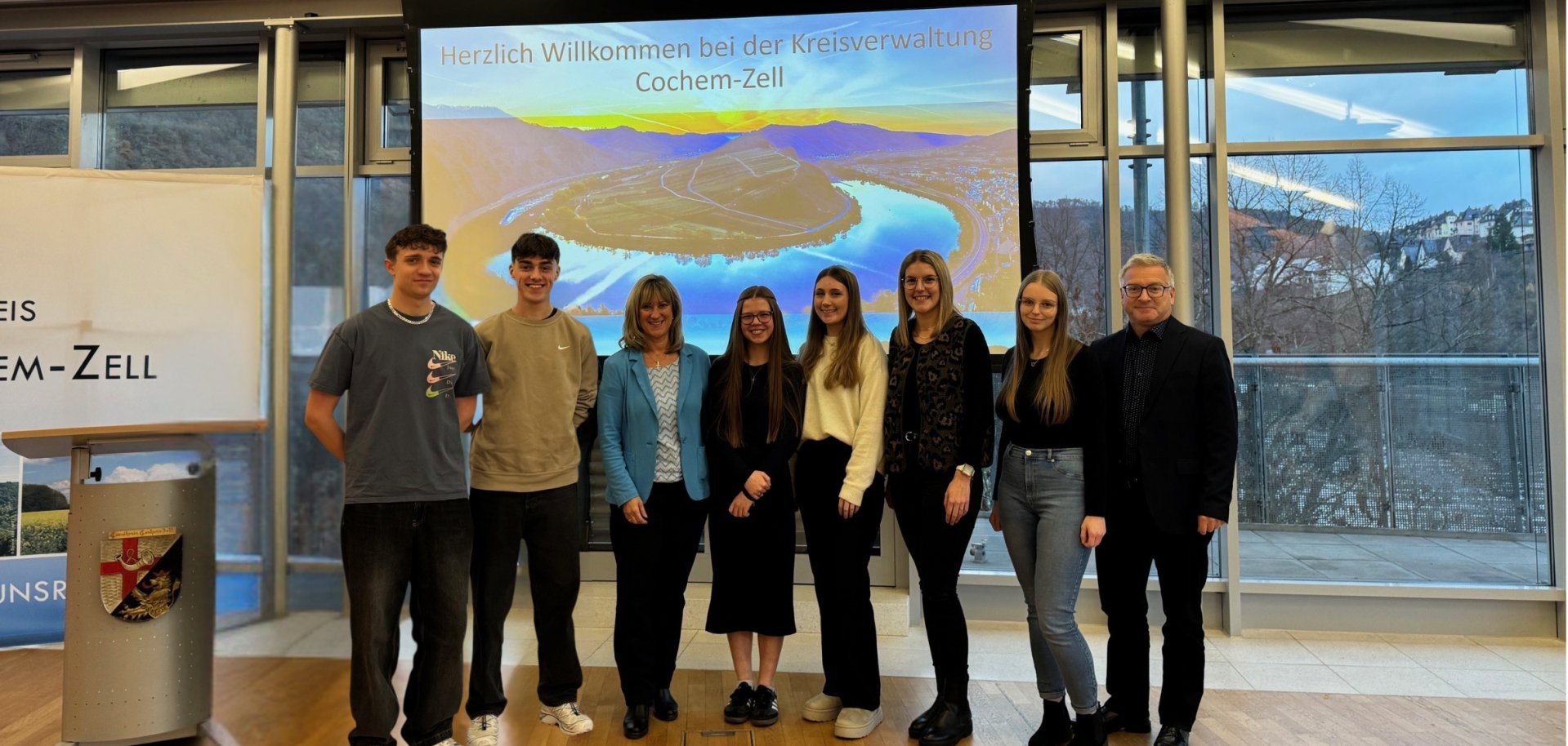 Zu sehen sind Andrej Stavburo, Joshua Ludwig, Landrätin Anke Beilstein, Jolaine Piellusch, Pia-Sophie Weyers, Hanna Schneiders, Jana Michelle Schmitz und Büroleiter Philipp Thönnes vor einer Kreis Cochem-Zell Leinwand.