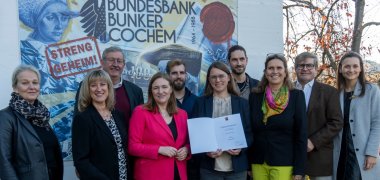 Eine Personengruppe steht draußen vor dem Eingang des  Bundesbankbunker in Cochem. v. l.: Roberta Castor (Beigeordnete VG Cochem), Landrätin Anke Beilstein, Stadtbürgermeister Walter Schmitz, Ministerin Katharina Binz, Marius Reuter, Antonia Mentel (Historikerin, Museumsleitung), Jean Reuter, Petra Reuter, Manf-red Reuter, Dominique Reuter
