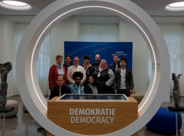 Zu sehen ist ein weißer Bogen. Auf dem Sockel steht "Demokratie, Democracy". Durch den Bogen schauen die Kursteilnehmer.