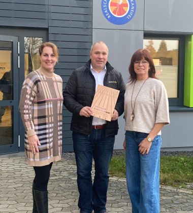 Übergabe der Siegerurkunde Zu sehen sind drei Personen die vor der Realschule plus Ulmen stehen. Konrektorin Nadine Fresia und Sportlehrerin Michaela Himberg nehmen eine Auszeichnung für den 1. Platz beim Westenergie Distanz-Duell durch Thomas Hau (Kommunalmanager Westenergie) entgegen.