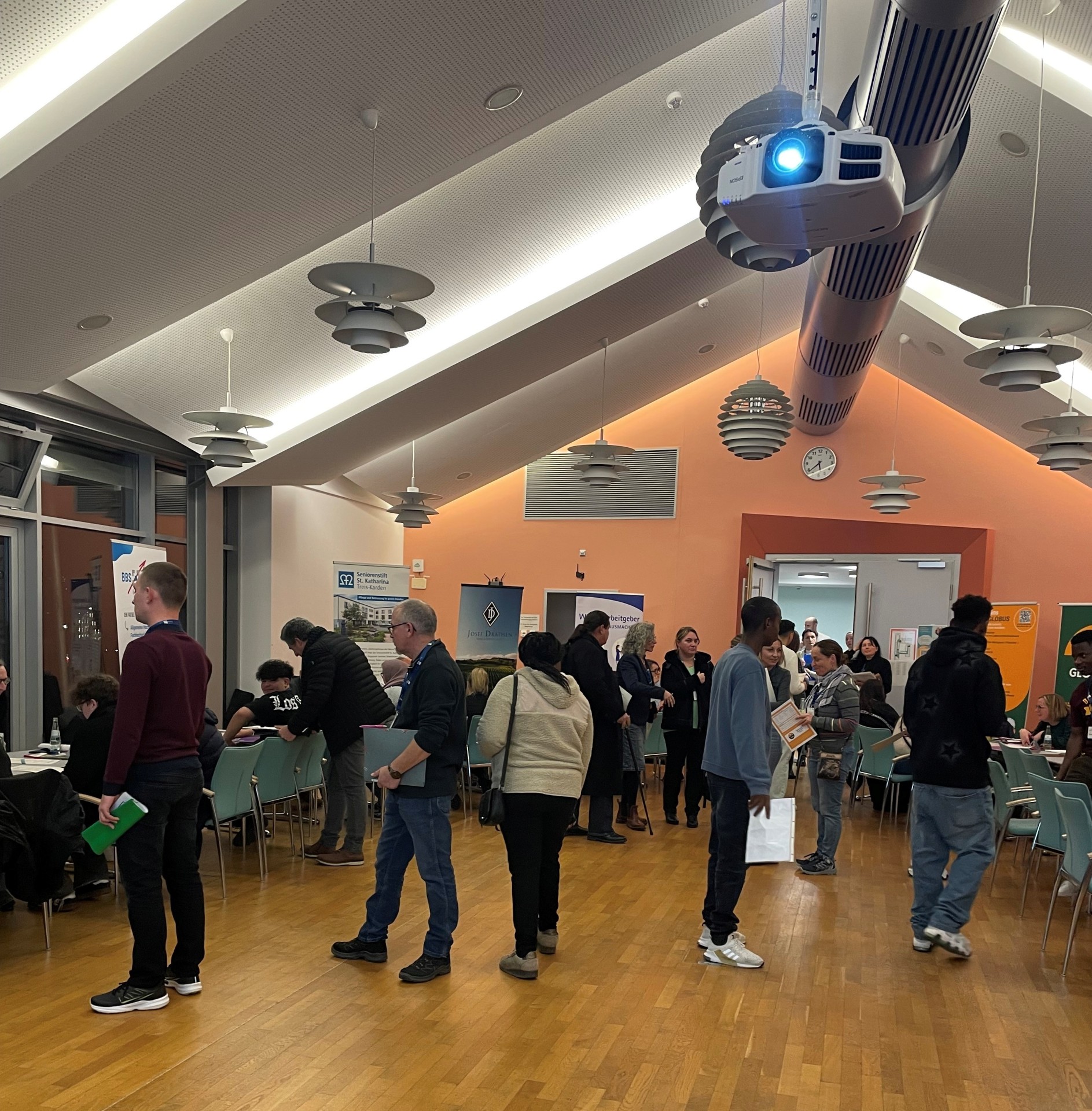 Zu sehen sind mehrere Personen, die sich frei im Raum der Jobmesse bewegen. Im Hintergrund sind Plakate der verschiedenen Berufe zu sehen.