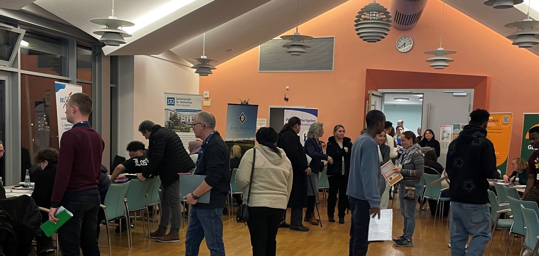 Zu sehen sind mehrere Personen, die sich frei im Raum der Jobmesse bewegen. Im Hintergrund sind Plakate der verschiedenen Berufe zu sehen.