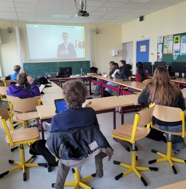 Zu sehen sind Schüler in einem Klassenzimmer. An der Tafel wird ein Videostream angezeigt.
