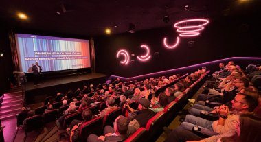 Zu sehen ist ein Kinosaal in dem eine Präsentation gehalten wird. Auf den Sessel sitzen verschiedene Personen.