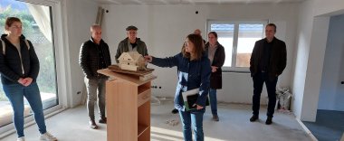 Zu sehen sind sieben Personen. Eine Frau von ihnen erklärt ein Modellhaus, welches die Sanierung alter Häuser darstellt.