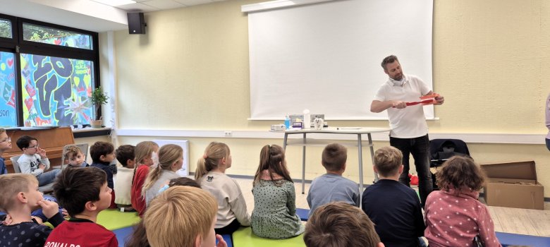 Grundschule Treis-Karden: Zahnvorsorge im Klassenzimmer Im Vordergrund des Bildes sitzen Schülerinnen und Schüler auf Sitzblöcken in einem Klassenraum und lauschen dem vor ihnen stehenden Zahnarzt Dr. Stein. Dieser demonstriert mithilfe eines überdimensionierten Modellgebisses und einer ebenso überdimensionierten Zahnbürste das korrekte Zähneputzen.