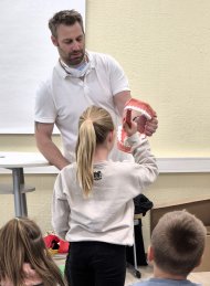 Grundschule Treis-Karden: Zahnvorsorge im Klassenzimmer Zahnarzt Dr. Stein hält ein überdimensioniertes menschliches Gebiss in den Händen. vor ihm steht eine Schülerin, die mit einer ebenso überdimensionierten Zahnbürse den Putzvorgang übt.