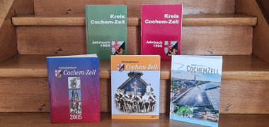Das Bild zeigt fünf Ausgaben des Heimatjahrbuchs Cochem-Zell, die auf einer Holztreppe dekorativ aufgestellt sind: in der oberen Reihe zwei Bücher, auf der Stufe darunter drei Bücher. Die Vorderseiten sind gut zu erkennen - ein Hinweis darauf, wie sich das Heimatjahrbuch über die Jahrzehte verändert hat.
