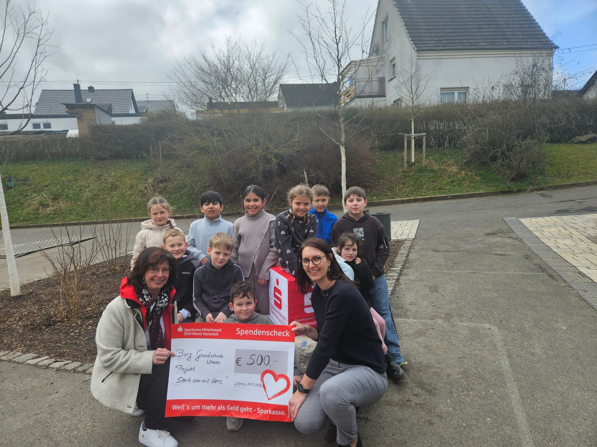 Scheckübergabe von Frau Bettina Kessler von der Sparkasse Mittelmosel an die Burg-Grundschule Ulmen, vertreten durch die Schulsozialarbeiterin Corinna Theisen, sowie Kinder, die am Training teilgenommen hatten. Die abgebildete Gruppe hat sich um den Scheck herum gestellt, der von den beiden Erwachsenen gehalten wird.Alle schauen gut gelaunt in die Kamera. 
