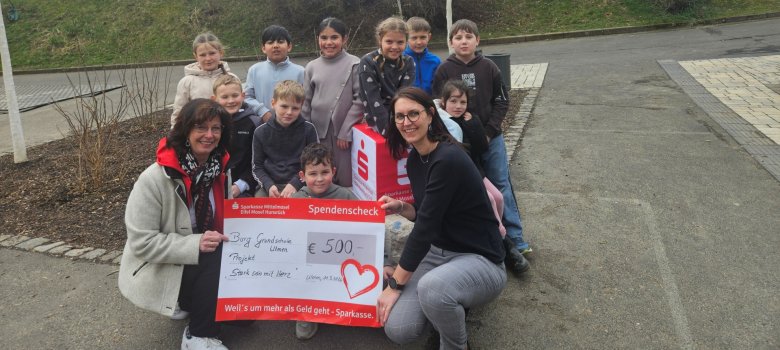 Scheckübergabe von Frau Bettina Kessler von der Sparkasse Mittelmosel an die Burg-Grundschule Ulmen, vertreten durch die Schulsozialarbeiterin Corinna Theisen, sowie Kinder, die am Training teilgenommen hatten. Die abgebildete Gruppe hat sich um den Scheck herum gestellt, der von den beiden Erwachsenen gehalten wird.Alle schauen gut gelaunt in die Kamera. 