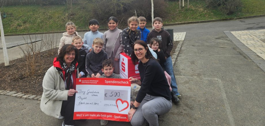 Scheckübergabe von Frau Bettina Kessler von der Sparkasse Mittelmosel an die Burg-Grundschule Ulmen, vertreten durch die Schulsozialarbeiterin Corinna Theisen, sowie Kinder, die am Training teilgenommen hatten. Die abgebildete Gruppe hat sich um den Scheck herum gestellt, der von den beiden Erwachsenen gehalten wird.Alle schauen gut gelaunt in die Kamera. 