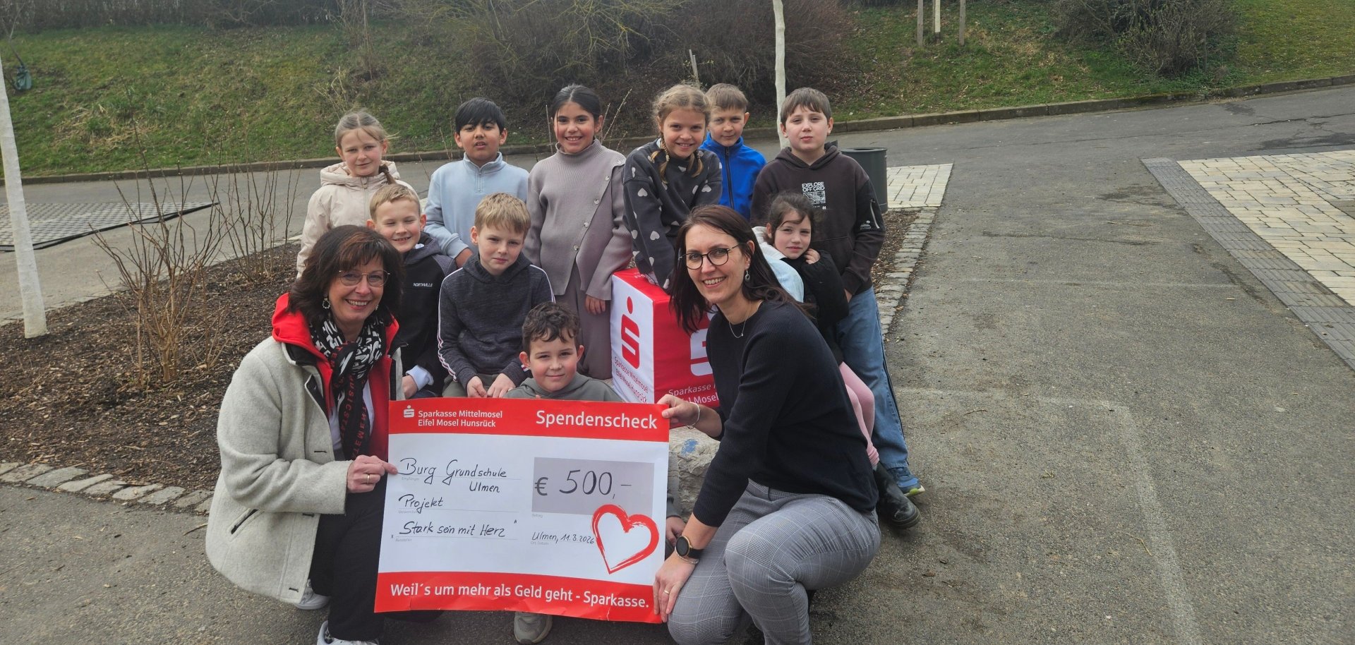 Scheckübergabe von Frau Bettina Kessler von der Sparkasse Mittelmosel an die Burg-Grundschule Ulmen, vertreten durch die Schulsozialarbeiterin Corinna Theisen, sowie Kinder, die am Training teilgenommen hatten. Die abgebildete Gruppe hat sich um den Scheck herum gestellt, der von den beiden Erwachsenen gehalten wird.Alle schauen gut gelaunt in die Kamera. 