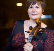 Zu sehen ist die Konzertpädagogin Katja Zakotnik. Sie trägt ein blau, rotes Kleid und in ihrer Hand hällt sie ein Cello.
