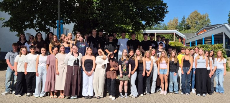 Großer Zuwachs an der FOS Kaisersesch Zu sehen sind Schülerinnen und Schüler der FOS Kaisersesch. Diese stehen in Reihen vor einem Baum. Einige sind vom Schatten des Baumes bedeckt, andere stehen in der Sonne.