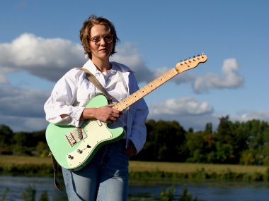 Das Foto zeigt die neue Gitarrenlehrerin an der Kreismusikschule Cochem-Zell: Lea Baciulis. Sie steht mit einer hellgrünen E-Gitarre im Vordergrund des Fotos. Die rechte Hand hält die Gitarre, die linke steckt in der Hosentasche. Sie trägt eine weiße, langärmlige Bluse und eine Jeans. Ihr Blick ist in die Kamera gerichtet. Im Hintergrund ist ein größeres Gewässer, dahinter ein Feld und ein Wald zu sehen. 