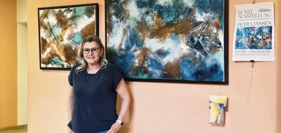 Kunst im Kreishaus Im Vordergrund steht die Künstlerin Frau Jansen. An der Wand befinden sich zwei Ihrer Künstwerke. Sie arbeitet mit der Pouring-Technik. Ihr Schwerpunkt ihrer Arbeiten liegt auf der abstarkten Malerei. Das eine Kunststück hat die Farben schwarz, dunkel blau, weiß, braun. Das zweite Bild hat die Farben braun, geld, weiß, türkis.