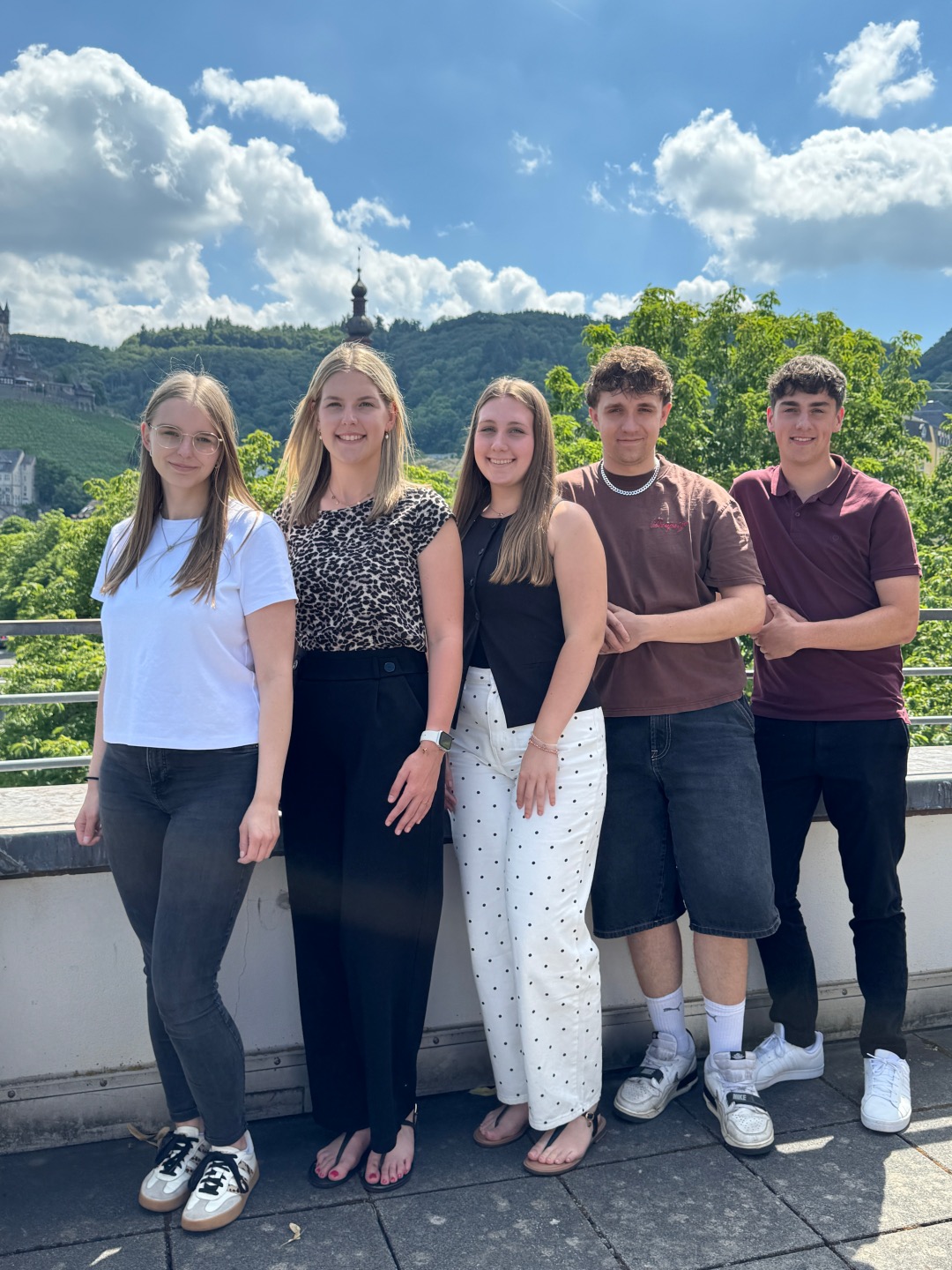 Zu sehen sind 5 Anwärter und Anwärterinnen des Kreisverwaltung Cochem-Zell. Von links nach rechts sind zu Beginn drei Frauen zu sehen und anschließend zwei Männer. Diese sind angemessen gekleidet. Sie befinden sich auf der Terasse der Kreisverwaltung. Im Hintergrund ist somit die die Natur erkennbar.