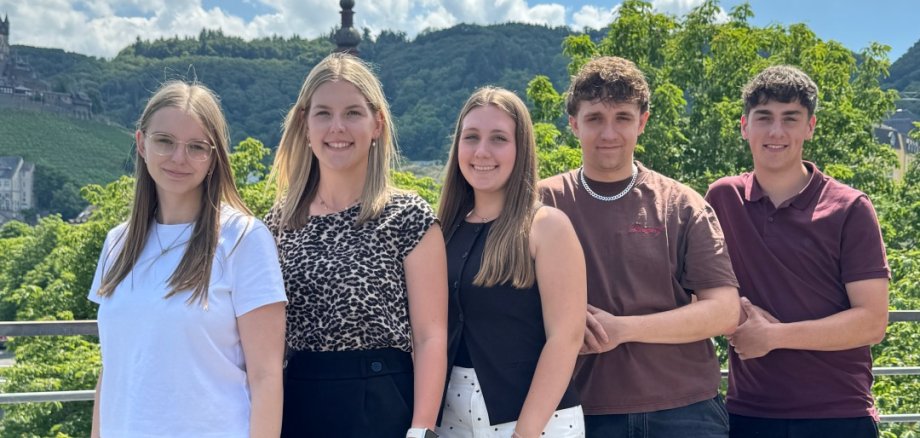 Neuer Lebensabschnitt: Wir begrüßen unsere neuen Nachwuchskräfte im 2. und 3. Einstieg-samt! Zu sehen sind 5 Anwärter und Anwärterinnen des Kreisverwaltung Cochem-Zell. Von links nach rechts sind zu Beginn drei Frauen zu sehen und anschließend zwei Männer. Diese sind angemessen gekleidet. Sie befinden sich auf der Terasse der Kreisverwaltung. Im Hintergrund ist somit die die Natur erkennbar.