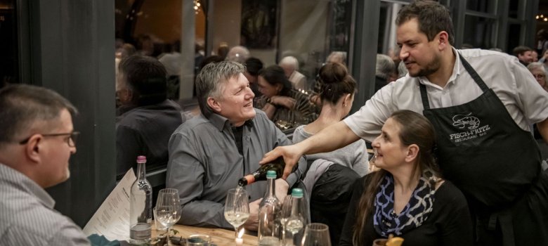 „Wein-Kulinarik-Reise“ Cochem-Zell geht 2027 in die nächste Runde zu sehen sind drei Personen, welche am Tisch sitzen. Sie sind fein gekleidet. Außerdem bekommen sie gerade Wein von einem Kellner eingeschenkt.