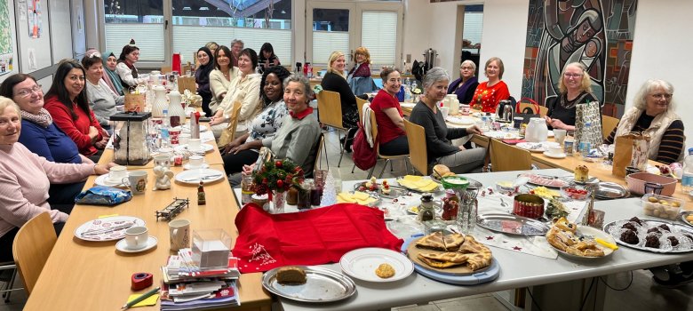 Rund 30 Frauen unterschiedlicher Herkunft sitzen beim letzten Internationalen Frauenfrühstück des Jahres 2025 zusammen. Sie sitzen an hufeisenförmig aufgstellten Tischen und lächeln in die Kamera. Vor ihnen steht Essgeschirr. Ganz im Vordergrund ist auf einem Tisch ein Buffet aus mitgebrachten Speisen zu sehen.