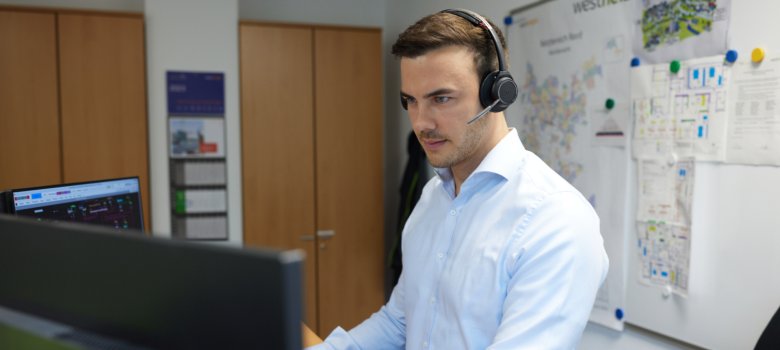 Zusehen ist ein Mann in einem Hemd, der vor seinem Computer sitzt mit einem Headset. Er bedient gerade die Störungshotline.