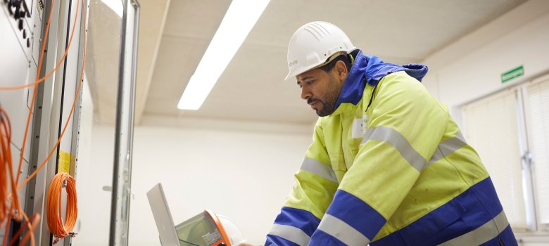 Zusehen ist ein Mann der Westnetz GmbH. Er hat eine Arbeitsjacke, sowie ein Helm an. Er befindet sich gerade im EDV-Raum, wo er auf einen Laptop schaut.