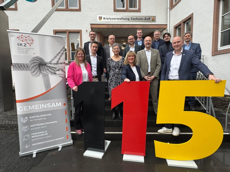 Ganz vorne auf dem Bild ist ein Austeller der 115 zusehen. Daneben steht das gemeinsame Roll Up des IKZ Verbundes. In der ersten Reihe steht v. l.n.r.: Landrätin Julia Gieseking (Landkreis Vulkaneifel), Landrätin Anke Beilstein (Landkreis Cochem-Zell) und Landrat Andreas Kruppert (Eifelkreis Bitburg-Prüm) und freuen sich über den Start des gemeinsamen 115-Servicecenters Eifel-Mosel-Hunsrück. Dahinter sind einige Personen von den beteiligten Kreisverwaltungen und Ministerien zusehen.