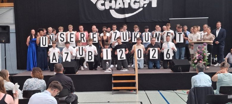 51 SchülerInnen an der Igs Zell feierlich verabschiedet Zu sehen sind Absolveten auf einer Bühne in festlicher Kleidung. Einige von ihnen halten Buchstaben hoch, welches im gesamten einen text ergibt welcher: "Unsere Zukunft Azubi 2026" lautet. Hinter ihnen steht auf einem Plakat: "AKI-Abschluss Dank CHATGPT"