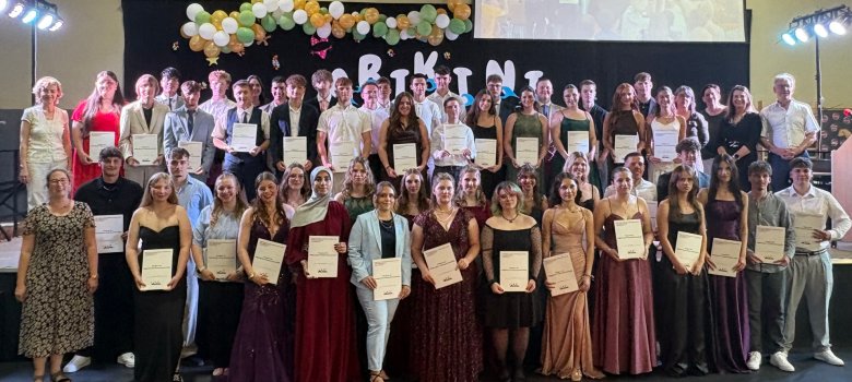 Abschluss öffnet viele Türen Zu sehen sind Schülerinnen und Schüler in festlicher Kleidung mit ihren Abschlusszeugnissen in der Hand. Diese stehen in Reihen auf einer Bühne. Im Hintergund befindet sich ein schwarzer Hintergrund mit dem Abschlussmotto "Abikini" drauf.