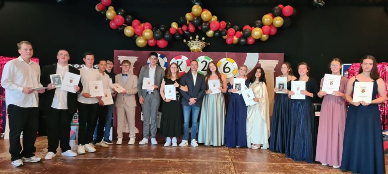 Abschlussfeier an der Realschule plus Vulkaneifel Zu sehen sind Schülerinnen und Schüler auf der Bühne in festlicher Kleidung. Diese halten ihre Abschlusszeugnisse der zehnten Klasse in der Hand.