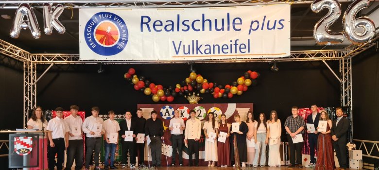 Abschlussfeier an der Realschule plus Vulkaneifel Es sind Schülerinnen und Schüler in festlicher Kleidung zu sehen. Diese stehen auf einer Bühne unter der Bunner: "Realschule plus Vulkaneifel". Es handelt sich hierbei um Neuntklässler, welche ihren Abschluss gemacht haben.