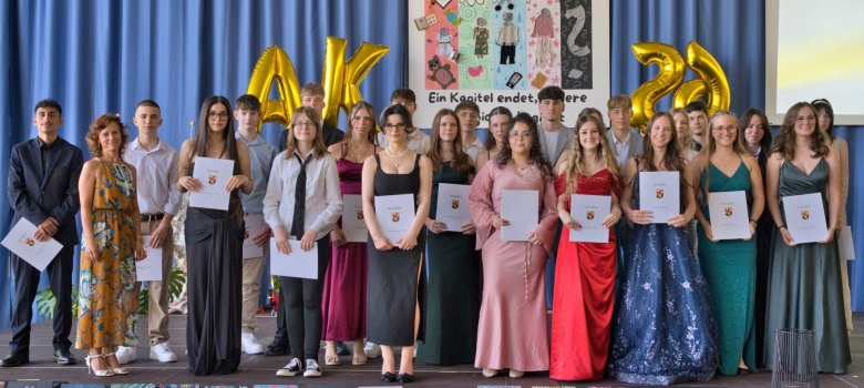 Die Abschlussklassen der Realschule plus Cochem schließen ihr Kapitel an der Schule ab Zu sehen sind Schülerinnen und Schüler der Neunten Klasse, welche nun ihren Abschluss haben. Diese sind festlich gekleidet und stehen in eihen auf der Bühne. Die Absolventen halten ihre Abschlusszeugnisse in der Hand. Vor ihnen befindet sich eine Austellung an gemalten Bildern, welche auf dem Boden der Bühne liegen. Im Hintergrund befindet sich ein Poster mit der Aufschrift: "Ein Kapitel endet, unsere Geschichte beginnt."
