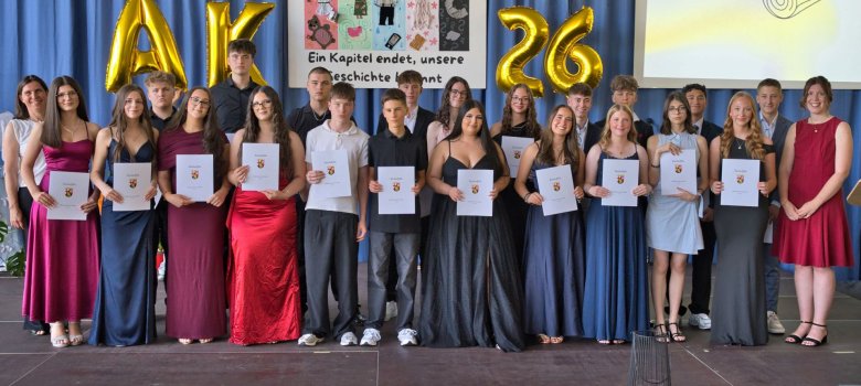 Die Abschlussklassen der Realschule plus Cochem schließen ihr Kapitel an der Schule ab Zu sehen sind Schülerinnen und Schüler der Neunten Klasse, welche nun ihren Abschluss haben. Diese sind festlich gekleidet und stehen in eihen auf der Bühne. Die Absolventen halten ihre Abschlusszeugnisse in der Hand. Vor ihnen befindet sich eine Austellung an gemalten Bildern, welche auf dem Boden der Bühne liegen. Im Hintergrund befindet sich ein Poster mit der Aufschrift: "Ein Kapitel endet, unsere Geschichte beginnt."