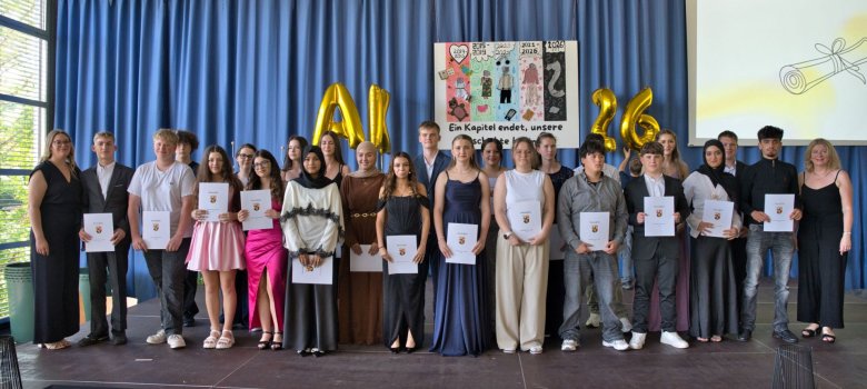Die Abschlussklassen der Realschule plus Cochem schließen ihr Kapitel an der Schule ab Zu sehen sind Schülerinnen und Schüler der Neunten Klasse, welche nun ihren Abschluss haben. Diese sind festlich gekleidet und stehen in eihen auf der Bühne. Die Absolventen halten ihre Abschlusszeugnisse in der Hand. Vor ihnen befindet sich eine Austellung an gemalten Bildern, welche auf dem Boden der Bühne liegen. Im Hintergrund befindet sich ein Poster mit der Aufschrift: "Ein Kapitel endet, unsere Geschichte beginnt."