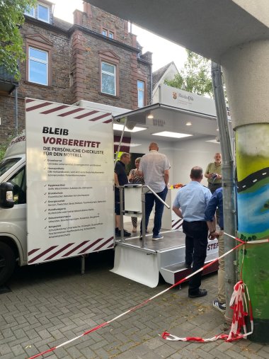 Bevölkerungsschutz im Fokus Zu sehen ist das Bleibbereit-Mobil, welches eine Informationsmöglichkeit zur persönlichen Nottfallvorsorge bietet. Es befindet sich im Innenhof der Kreisverwaltung Cochem-Zell. Landrätin Anke Beilstein sowie weitere Personen sind am Infostand im Innenraum des Mobils zu sehen.
