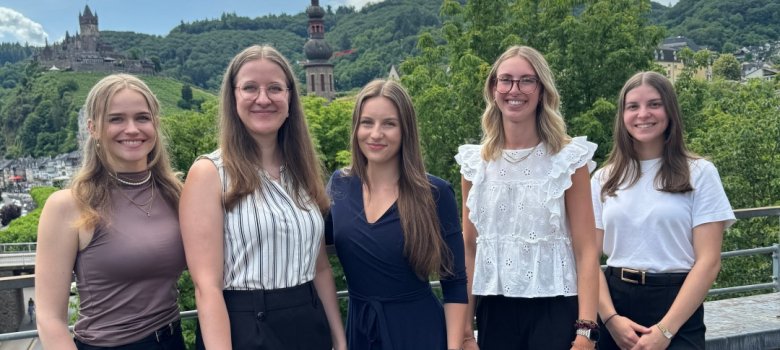 Zu sehen sind 5 Damen. Diese stehen in feiner Kleidung auf der Terasse eines Gebäudes. Im Hintergrund ist die Burg Cochem zu sehen.