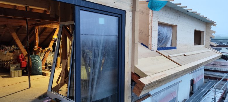 Das Foto zeigt eine Baustelle: Es ist ein Dachstuhl eines Wohnhauses zu sehen - durch ein großes Fenster sieht man innen das Gebälk des alten Dachstuhls, außen sind Dämmung und neue Gauben zu sehen, das Dach ist noch nicht gedeckt.
