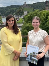 Landkreis Cochem-Zell vergibt zum dritten Mal Medizinstipendien Das Bild zeigt Gesundheitsmanagerin Julia Wilde (links) und Stipendiatin Alina Kühn (rechts). im Hintergrund sieht man Bäume und einige Gebäude. Alina Kühn hält eine Mappe in der Hand.