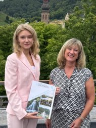Landkreis Cochem-Zell vergibt zum dritten Mal Medizinstipendien Das Bild zeigt Landrätin Anke Beilstein (rechts) und Stipendiatin Anna Mayers (links). im Hintergrund sieht man Bäume und einige Gebäude. Anna Mayers hält eine Mappe in der Hand.