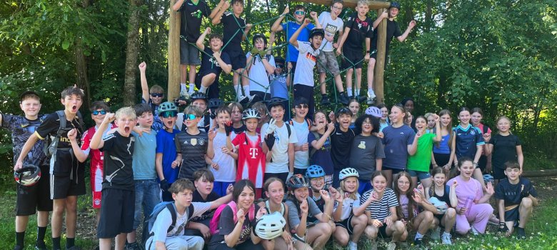 Mountainbike-Tag für die drei Sportprofilklassen der GOS Cochem Zu sehen sind Schüler und Schülerinnen der sechsten Klasse. Diese stehen teilweise in Reihen aufgereit nebeneinander mit der Faust nach oben. Einige knien vor den aufgereiten Schülern. Einige hängen im Balance Parcour Netz. Sie befinden sich auf einer Wiese und hinter ihnen sind Bäume.