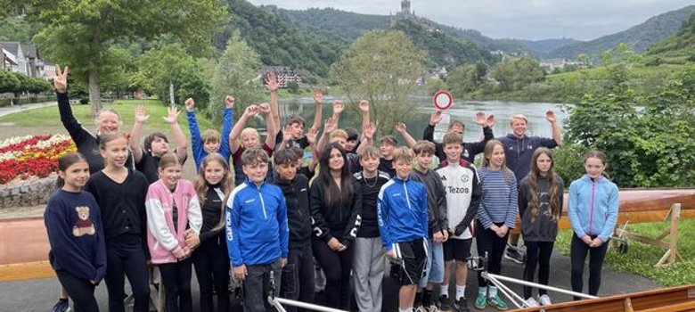 Sportprofilklassen der GOS Cochem zu Gast beim Cochemer Ruderverein Zu sehen sind Kinder jungen Alters, welche in Reihen zwischen Ruderbooten stehen. Einige von ihnen haben die Hände gehoben. Im Hintergrund befindet sich die Mosel