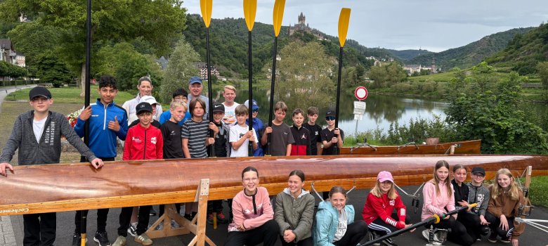 Sportprofilklassen der GOS Cochem zu Gast beim Cochemer Ruderverein Zu sehen sind Kinder jungen Alters. Die Mädchen dieser Schulklasse knien vor dem Ruderboot. Die Jungs stehen in Reihen hinter dem Boot und halten Paddel nach oben. Dahinter stehen noch 3 Lehrer. Im Hintergrund befindet sich die Mosel.