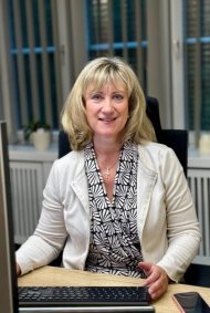 Landrätin Anke Beilstein sitzt an ihrem Schreibtisch. Sie schaut in die Kamera und lächelt. Vor ihr sind ihr Computerbildschirm, der Laptop und ihr Handy zu sehen.