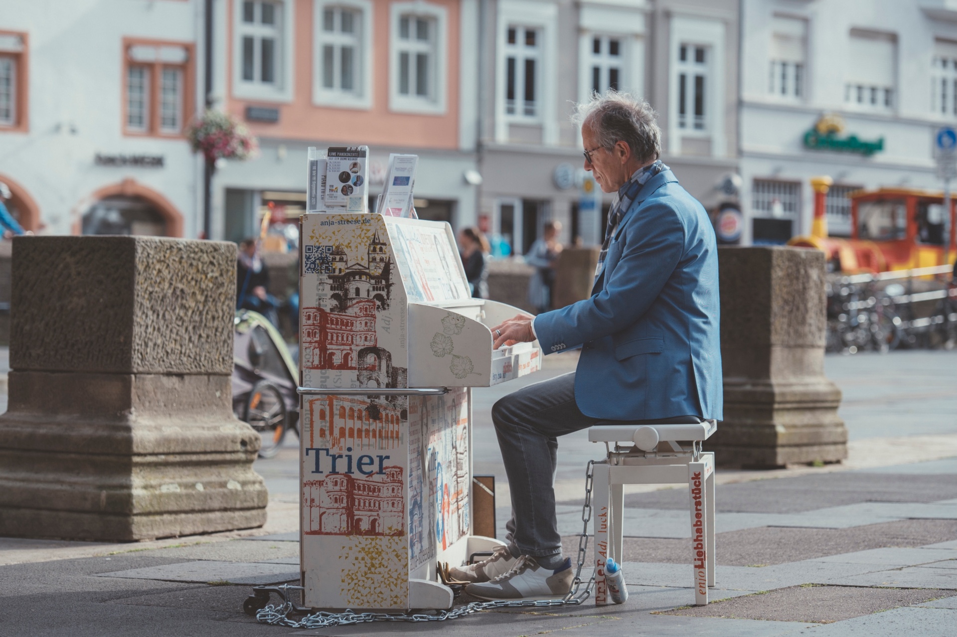 Zu sehen ist ein Mann, der an einem Klavier sitzt und spielt. Das Klavier steht dabei mitten auf einem Marktplatz.